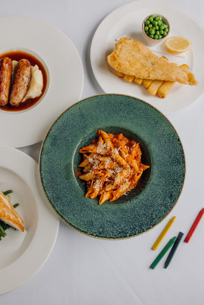 Small plates of pasta with cheese, fish and chips, sausage and mash, chicken and veg laid on a table with crayons
