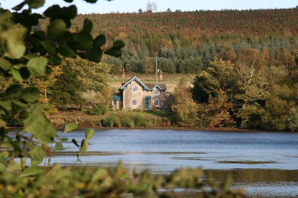A large lake with reflections on, a house sat in the middle with woodlands behind.