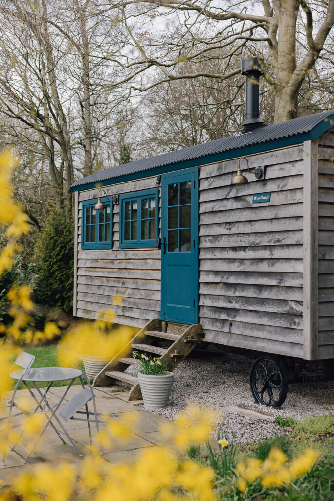 Outside of a shepherds hut with blue windows and doors, patio with bistro set for two