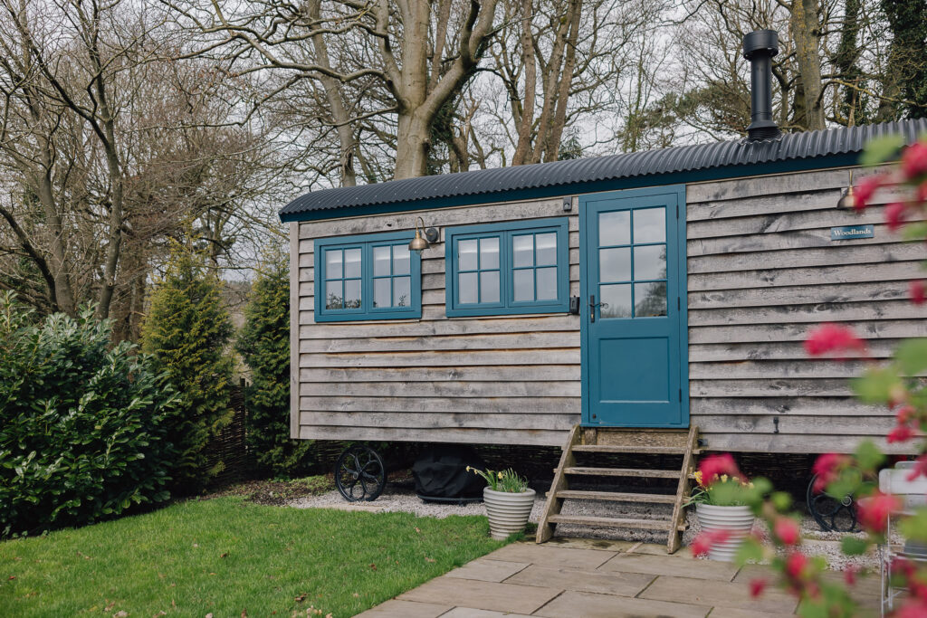 Outside of a shepherds hut with blue windows and doors, patio with bistro set for two